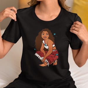 May include: Black t-shirt with a cartoon illustration of a Polynesian woman with long brown hair wearing a red and white patterned skirt and a white top. The woman is sitting with her legs crossed and has a necklace on. There are blue stars around her.