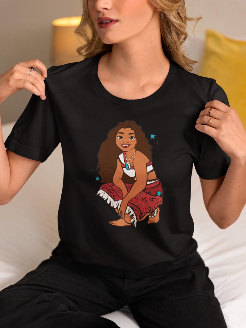 May include: Black t-shirt with a cartoon illustration of a Polynesian woman with long brown hair wearing a red and white patterned skirt and a white top. The woman is sitting with her legs crossed and has a necklace on. There are blue stars around her.