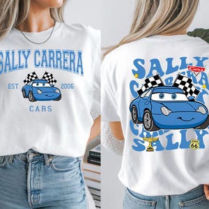 Pode incluir: Camiseta branca com um carro de desenho animado azul, bandeiras quadriculadas e o texto "SALLY CARRERA" e "CARS". O design frontal inclui "EST. 2006". O design traseiro repete o gr&aacute;fico do carro e o texto.