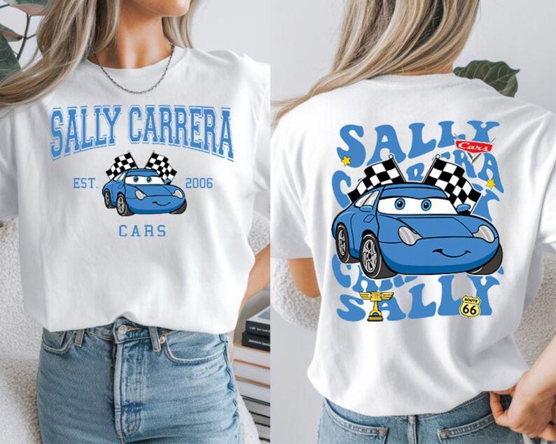 Pode incluir: Camiseta branca com um carro de desenho animado azul, bandeiras quadriculadas e o texto "SALLY CARRERA" e "CARS". O design frontal inclui "EST. 2006". O design traseiro repete o gr&aacute;fico do carro e o texto.