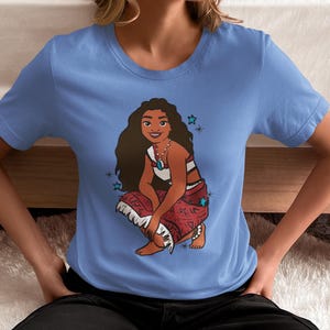 May include: A light blue t-shirt with a cartoon image of a woman with long brown hair wearing a red and white patterned skirt. The woman is sitting with her legs crossed and has a blue necklace with a turquoise pendant. There are blue stars around the woman.