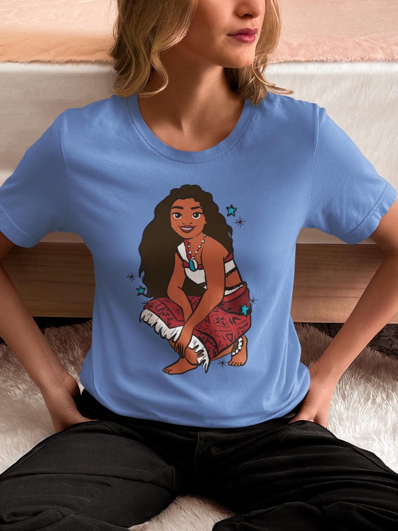 May include: A light blue t-shirt with a cartoon image of a woman with long brown hair wearing a red and white patterned skirt. The woman is sitting with her legs crossed and has a blue necklace with a turquoise pendant. There are blue stars around the woman.