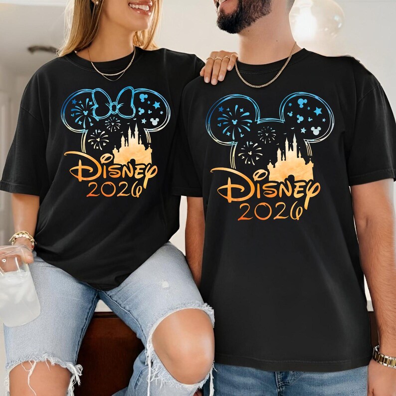 May include: Two black t-shirts featuring a graphic of a Mickey Mouse ear silhouette filled with fireworks, stars, and a castle. The design includes the text Disney 2026 in an orange gradient font. The shirts are worn with light blue denim jeans.