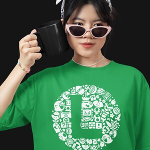 May include: Green t-shirt with a white graphic design featuring various video game characters and icons arranged in a circular pattern. The shirt is made of a soft material. A black mug is held in the hand.