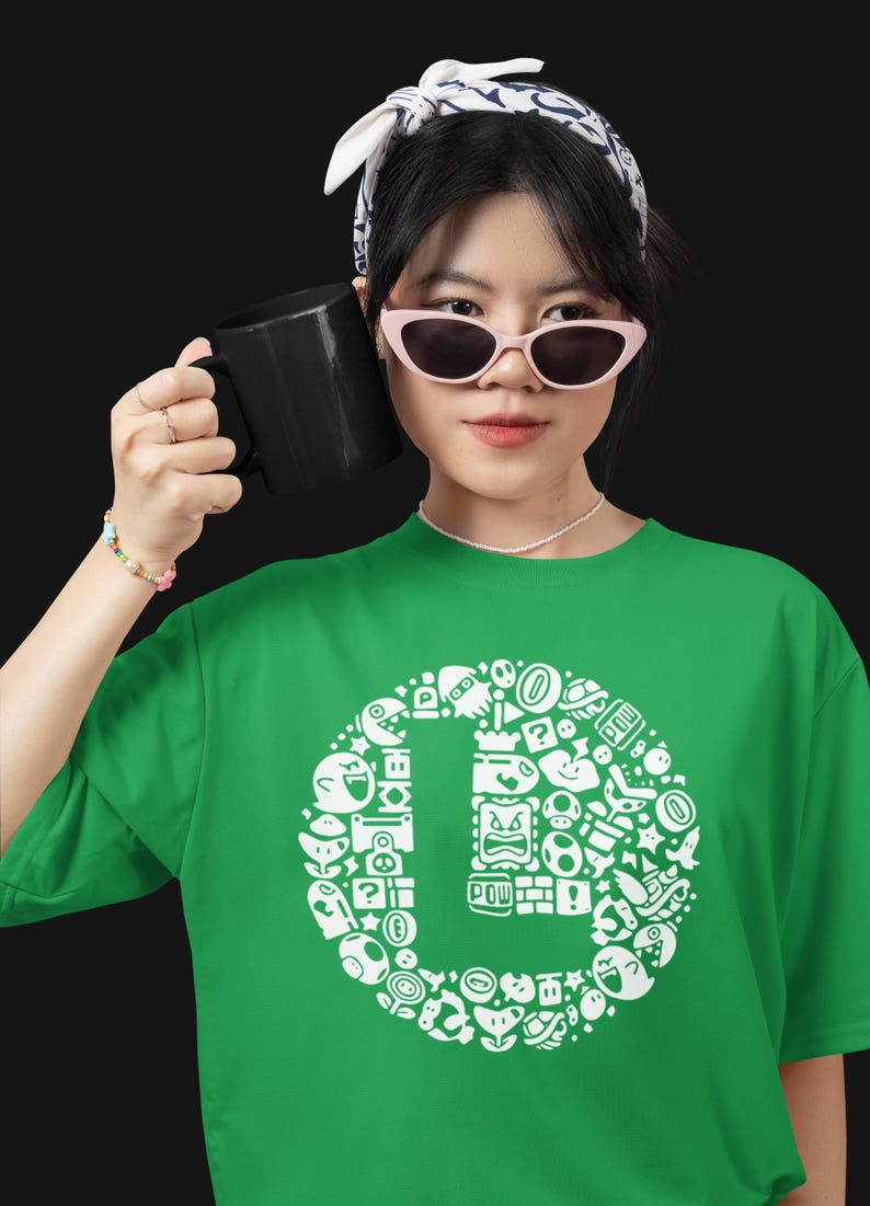 May include: Green t-shirt with a white graphic design featuring various video game characters and icons arranged in a circular pattern. The shirt is made of a soft material. A black mug is held in the hand.