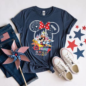 May include: A navy blue t-shirt featuring a white outline of Mickey Mouse ears containing a castle, fireworks, and Disney characters. Below the graphic, the word Disney is printed in red, followed by the text 2026 with character-themed numbers.
