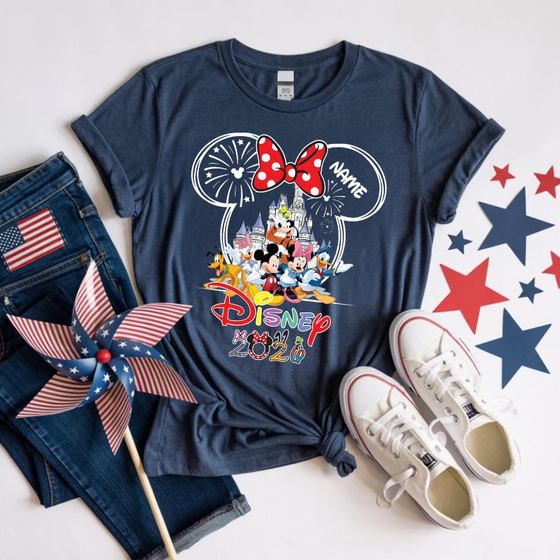 May include: A navy blue t-shirt featuring a white outline of Mickey Mouse ears containing a castle, fireworks, and Disney characters. Below the graphic, the word Disney is printed in red, followed by the text 2026 with character-themed numbers.