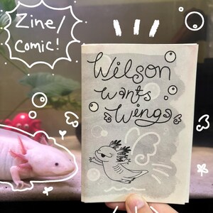 May include: A zine or comic book titled "Wilson Wants Wings" is held in front of a white axolotl in an aquarium. The cover features a cartoon axolotl with wings. The text "Zine/Comic!" is written in a speech bubble.