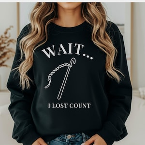 May include: Black crew neck sweatshirt with the white text "WAIT... I LOST COUNT". A white graphic of a crochet hook and chain is also featured. The sweatshirt is a casual style.
