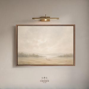 Può includere: Un dipinto di paesaggio rettangolare in una cornice di legno, con una tavolozza di colori tenui beige, beige chiaro e grigio. L'opera d'arte raffigura un campo, alberi lontani e un cielo nuvoloso. Una lampada per quadri in ottone è montata sopra la cornice. Il dipinto è etichettato "CHOSEN BY HIM".