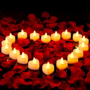 May include: Heart-shaped LED candles with a warm, yellow glow are arranged on a bed of red rose petals. The candles are white and create a romantic ambiance. The background is dark, emphasizing the candles and petals.