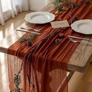 May include: A table setting with a burnt orange table runner. White plates with decorative edges, silverware, pinecones, and greenery are arranged on a wooden table. A card reads "FestFiesta". The runner has a textured weave.