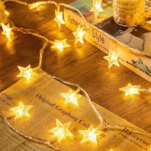 Op de afbeelding: Een decoratieve lichtslinger met warmwitte stervormige lampjes. De lampjes zijn op een transparante draad en liggen op een houten oppervlak en een vintage krant. Op de achtergrond ligt een boek met de tekst "American Style".