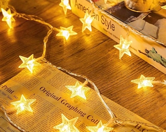 May include: A string of warm white star-shaped fairy lights. The lights are arranged on a thin, clear wire, and are scattered across a wooden surface and vintage newspaper. A book with the text "American Style" is in the background.