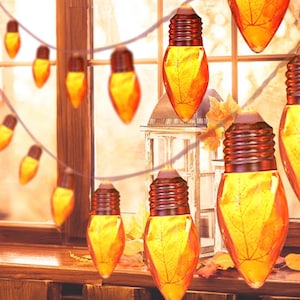 May include: A string of decorative light bulbs shaped like teardrops, each containing a preserved orange and yellow autumn leaf. The bulbs are connected by a thin wire, and the glass surfaces feature a warm, glowing amber hue with visible leaf veins.