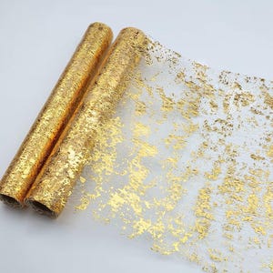 May include: Two rolls of gold-colored, decorative fabric. The fabric has a transparent base with a gold foil overlay, creating a textured, metallic effect. The rolls are tightly wound, and the fabric is partially unrolled, revealing the gold leaf design.