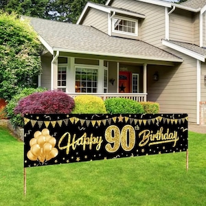 May include: A black banner with gold text and accents that reads "Happy 90th Birthday." The banner features gold balloons and star accents. It is displayed on a green lawn in front of a house.
