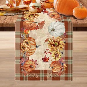 May include: A rectangular table runner featuring a beige centre panel decorated with orange pumpkins, yellow sunflowers, and red maple leaves. The border consists of a brown, beige, and green plaid pattern framing the autumn-themed design.