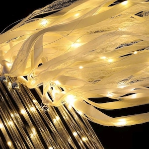 May include: A decorative light fixture featuring cascading cream-colored ribbons and delicate lace trim intertwined with thin, glowing LED string lights. The design incorporates multiple vertical strands of warm, illuminated lights that create a soft glow.
