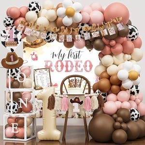 Puede incluir: Decoración de fiesta de primer cumpleaños con temática de rodeo. La escena incluye un arco de globos en tonos marrón, rosa y blanco, globos con estampado de vaca y una pancarta con fotos. Las palabras "My first Rodeo" se muestran de forma destacada.