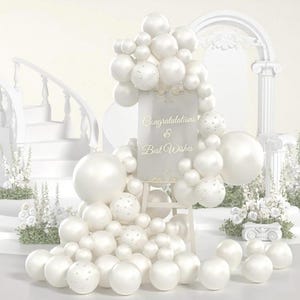 May include: A white balloon arch and display with the words "Congratulations & Best Wishes" on a sign. The display includes a staircase, columns, and floral arrangements, all in white, creating a celebratory atmosphere.
