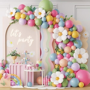 May include: A party decoration with a pastel colour scheme, including balloons, flowers, and a cake. The backdrop features the words "let's party" in white script. The arrangement includes a rainbow, cupcakes, and a tiered dessert stand.