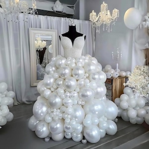 May include: A mannequin wearing a white balloon dress with a pearl-covered bodice. The dress is made of white balloons of different sizes. The background features white curtains, a mirror, and chandeliers, creating a celebratory setting.