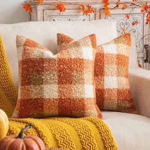 May include: Two square throw pillows featuring a textured, fuzzy sherpa-style fabric with a large buffalo check pattern in shades of burnt orange, cream, and tan. The pillows are arranged side-by-side, showcasing a soft, plush, and tactile surface.