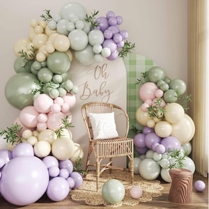 May include: A baby shower scene with pastel-colored balloons forming arches and clusters. A wicker chair with a white pillow sits in front of a backdrop with the words "Oh Baby." Decorative greenery and a wooden stump add to the festive decor.