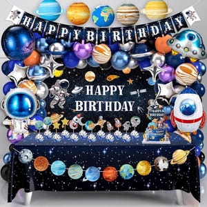 May include: A space-themed birthday party setup with a dark blue tablecloth adorned with planets. The scene includes balloons, a banner that says "Happy Birthday", and cake toppers. The color scheme features blues, purples, and silver.