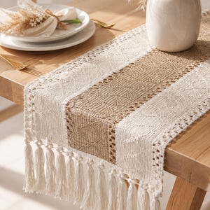 May include: A rectangular table runner featuring alternating vertical stripes of cream-colored woven fabric and a central tan textured panel. The edges are finished with a decorative crochet border and long, hanging cream-colored tassels.