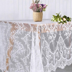 May include: A white lace tablecloth draped over a wooden table. The intricate floral pattern of the lace is visible. A wicker basket with flowers and a small bouquet of flowers are on the table.