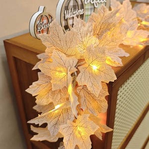 May include: A string of artificial maple leaves with integrated warm yellow LED lights. The leaves are light beige with visible dark veining. Behind the leaves, two circular wooden signs are visible, one featuring the text Bless and the other Welcome.