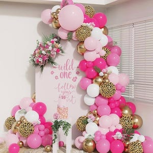 Può includere: Decorazione per feste con un arco di palloncini rosa e oro con accenti leopardati. L'esposizione comprende un cartello con le parole "wild one follow your art" e una torta su un piedistallo.