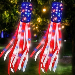 May include: Two patriotic windsock decorations. Each has a blue cylinder top with white stars and red, white, and blue ribbons. The ribbons have embedded lights. The background is a blurred outdoor scene.
