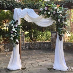 Op de afbeelding: Een houten trouwboog versierd met witte stoffen draperieën en bloemstukken. De boog is versierd met witte, roze en bordeauxrode bloemen en groen. De setting is buiten, met een stenen muur en bomen op de achtergrond.
