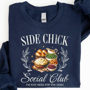 May include: Navy blue sweatshirt with the text "SIDE CHICK Social Club" and the phrase "I'M JUST HERE FOR THE SIDES". The graphic features illustrations of various side dishes, including rolls, mashed potatoes, and cranberry sauce.