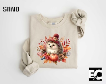 May include: Sand-colored sweatshirt featuring a watercolor illustration of a hedgehog wearing a red hat, surrounded by autumn leaves, berries, and a pumpkin. The design is centered on the front of the long-sleeved top.