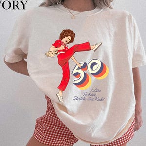 A white t-shirt with a graphic design depicting a woman in red pants and a red and white checkered skirt, along with the text "I like to kick, stretch, and kick!"