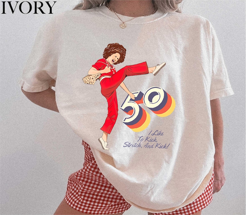 A white t-shirt with a graphic design depicting a woman in red pants and a red and white checkered skirt, along with the text "I like to kick, stretch, and kick!"
