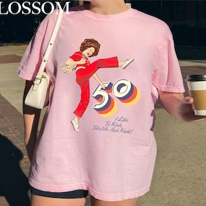 A person wearing a pink t-shirt with a graphic design of a woman in red pants and a white shoe, holding a coffee cup.