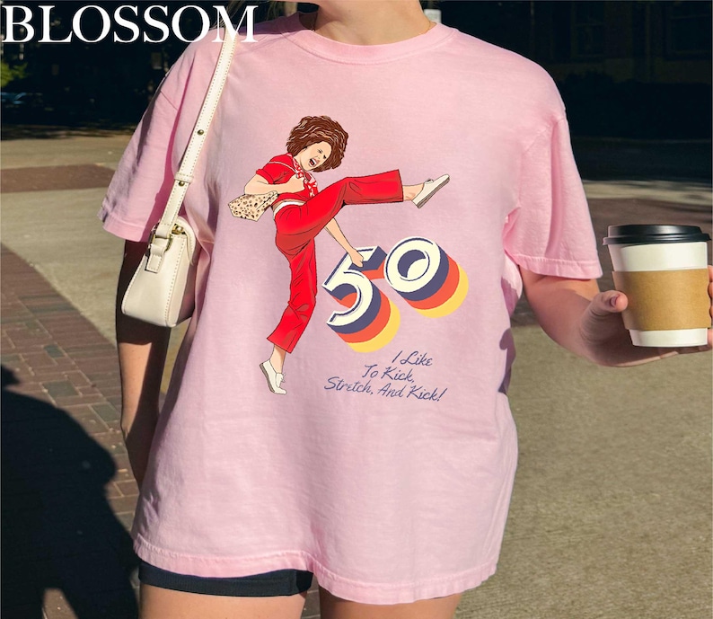 A person wearing a pink t-shirt with a graphic design of a woman in red pants and a white shoe, holding a coffee cup.