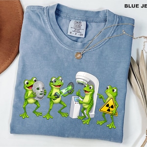 May include: A blue jean colored t-shirt featuring a graphic of four green frogs. One frog holds a silver mask, another uses a laser device, a third operates a white medical machine, and the fourth holds a yellow radiation warning sign.