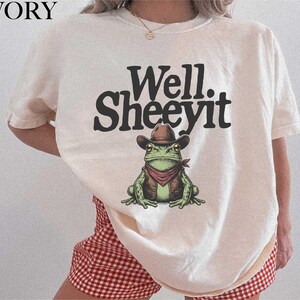 May include: Ivory-colored t-shirt with the text "Well. Sheeeyit" in black. The shirt features a cartoon frog wearing a cowboy hat, vest, and red bandana. The frog is green with brown and red accents. The shirt is part of a clothing collection.