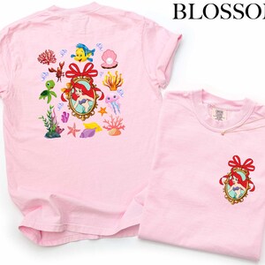 a pink t-shirt with a Disney-themed design, including a mermaid, fish, and other sea creatures. The t-shirt is displayed alongside a smaller version of the same t-shirt, which is folded and placed on a white background.