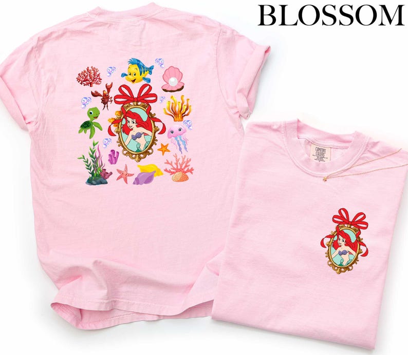 a pink t-shirt with a Disney-themed design, including a mermaid, fish, and other sea creatures. The t-shirt is displayed alongside a smaller version of the same t-shirt, which is folded and placed on a white background.