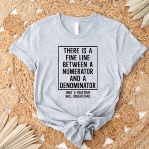 May include: A gray t-shirt with a black box containing the text "THERE IS A FINE LINE BETWEEN A NUMERATOR AND A DENOMINATOR ONLY A FRACTION WILL UNDERSTAND".