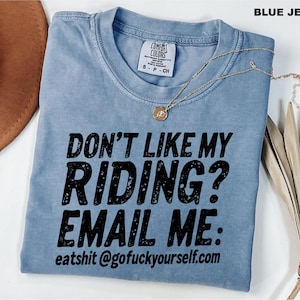 May include: A blue jean colored t-shirt with black text that reads: DON'T LIKE MY RIDING? EMAIL ME: eatshit@gofuckyourself.com. The shirt features a crew neckline, short sleeves, and a Comfort Colors brand label on the inside of the collar.