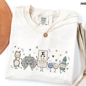 May include: An ivory t-shirt with a graphic print featuring cartoon-style eye care items including glasses, a phoropter, an eye chart with the letter E at the top, a contact lens case, and an eye drop bottle, all with smiling faces and arms.