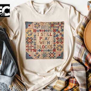 May include: Cream-colored t-shirt featuring a colorful patchwork design with the text "I STILL PLAY WITH BLOCKS." The patchwork design includes red, blue, and yellow geometric shapes. The shirt is styled with a denim jacket and a plaid scarf.